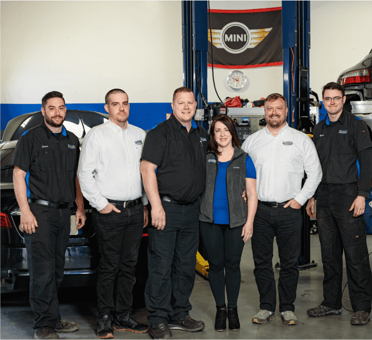 6 people huddle together and smile, looking at the camera, inside European Service Center’s auto repair shop.